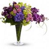 Bountiful blooms including purple hydrangea, green cymbidium orchids, purple mokara orchids, lavender roses, green viburnum, green button spray mums and purple stock are elegantly mixed with greens such as bupleurum, calathea leaves, philodendron leaves and lily grass in a tall, footed flare vase.
